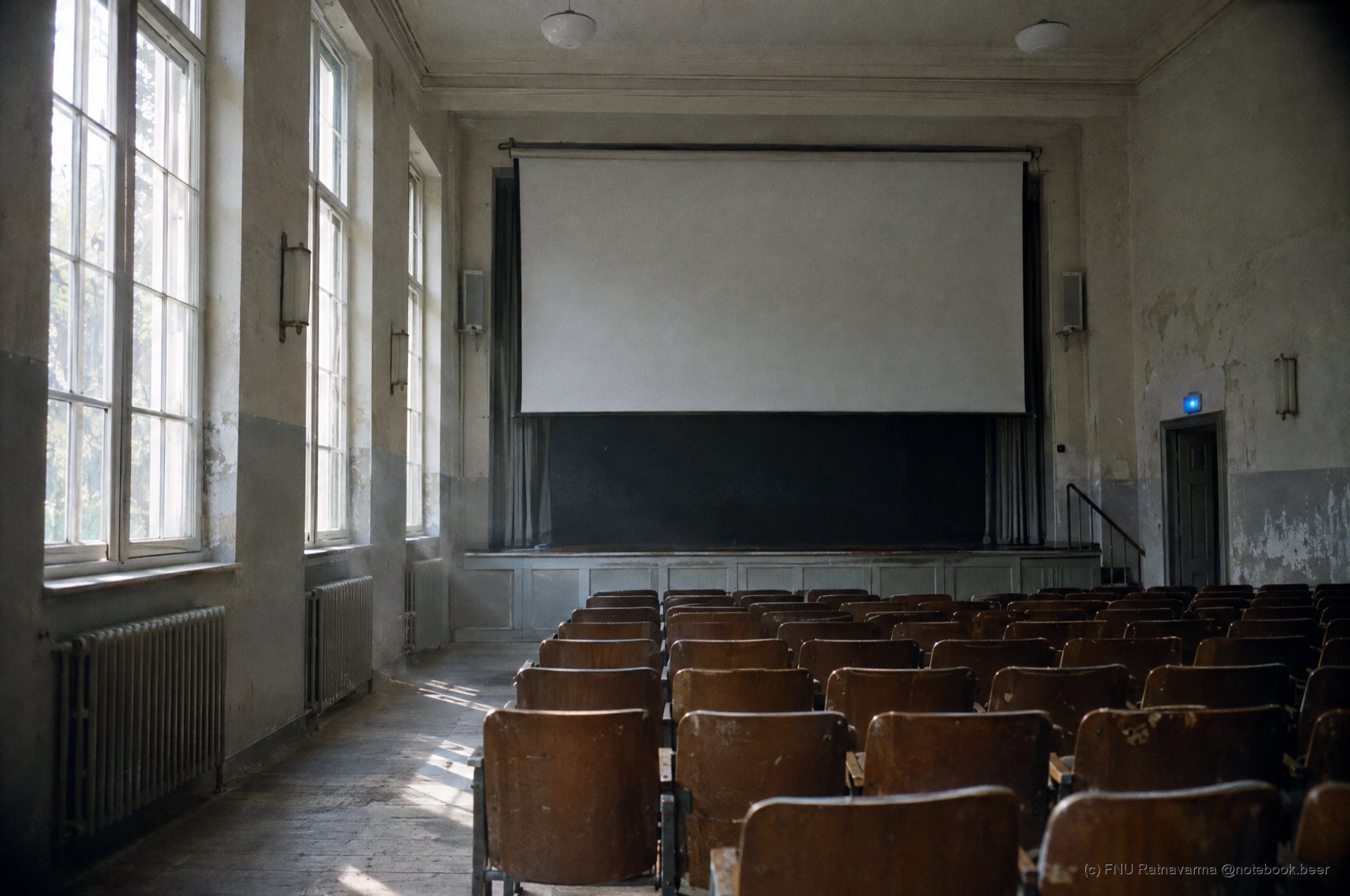 An old lecture hall in morning light, rows of wooden seats facing a blank projection screen.
